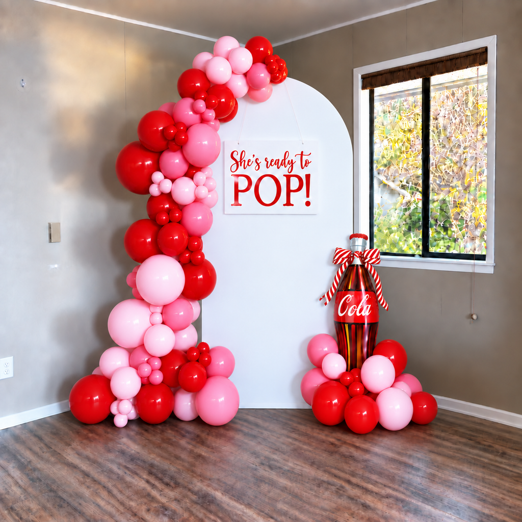 A pink and red balloon decoration setup in a room, featuring a large arch on the left and a cluster next to a white arched backdrop with the text 'She's ready to POP!' and a Coca-Cola bottle decoration.
