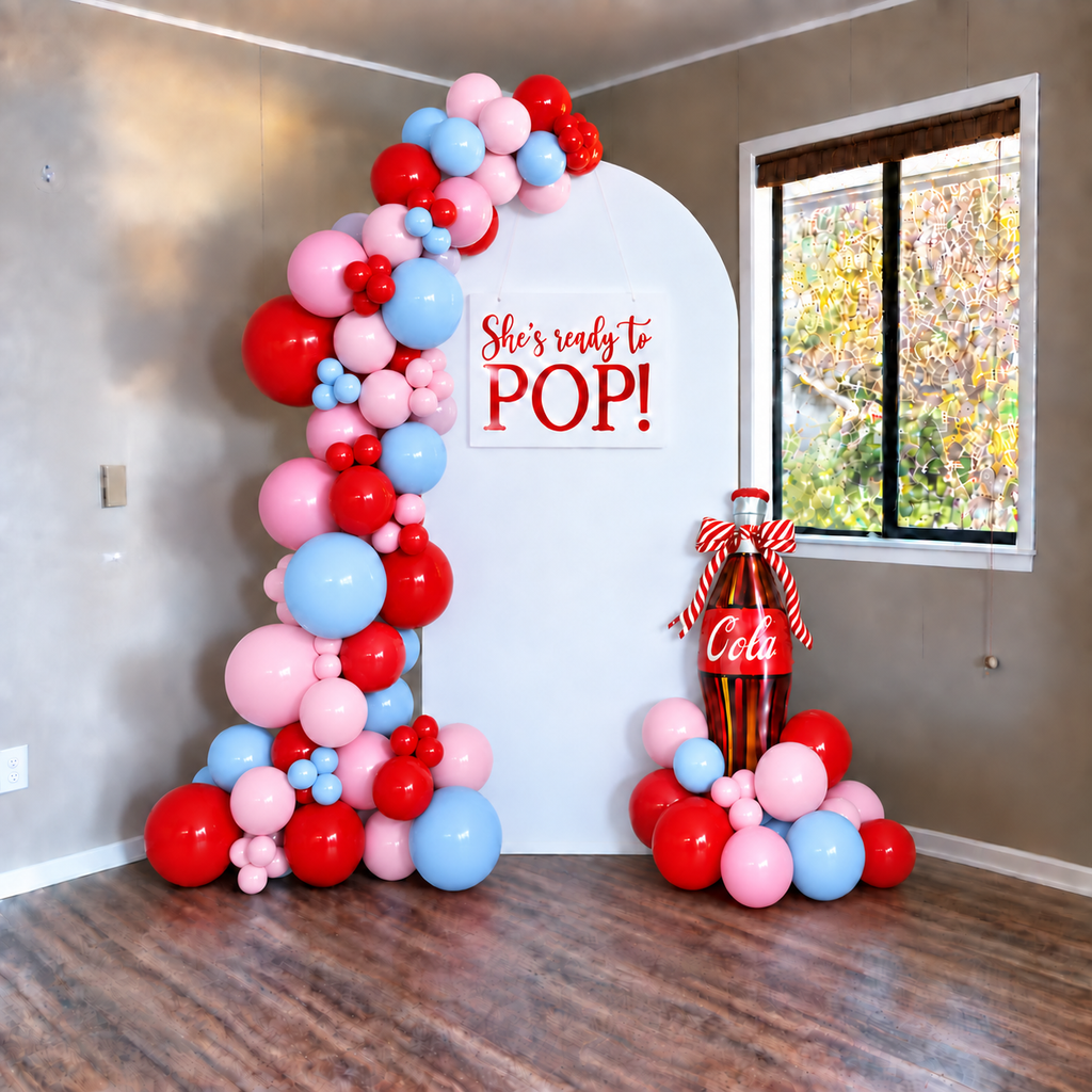 A red, pink, and light blue balloon garland arch and matching balloon cluster set up in a room, with a white arched backdrop sign reading 'She's ready to POP!' and a large Coca-Cola bottle decoration on the floor.